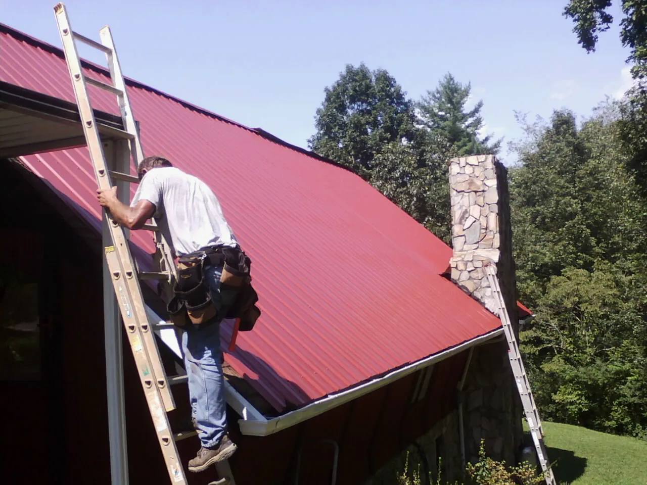 Roofer on a ladder beside a red metal roof and stone chimney — Soffit & Fascia Repair by Roofing Brothers in Rockdale